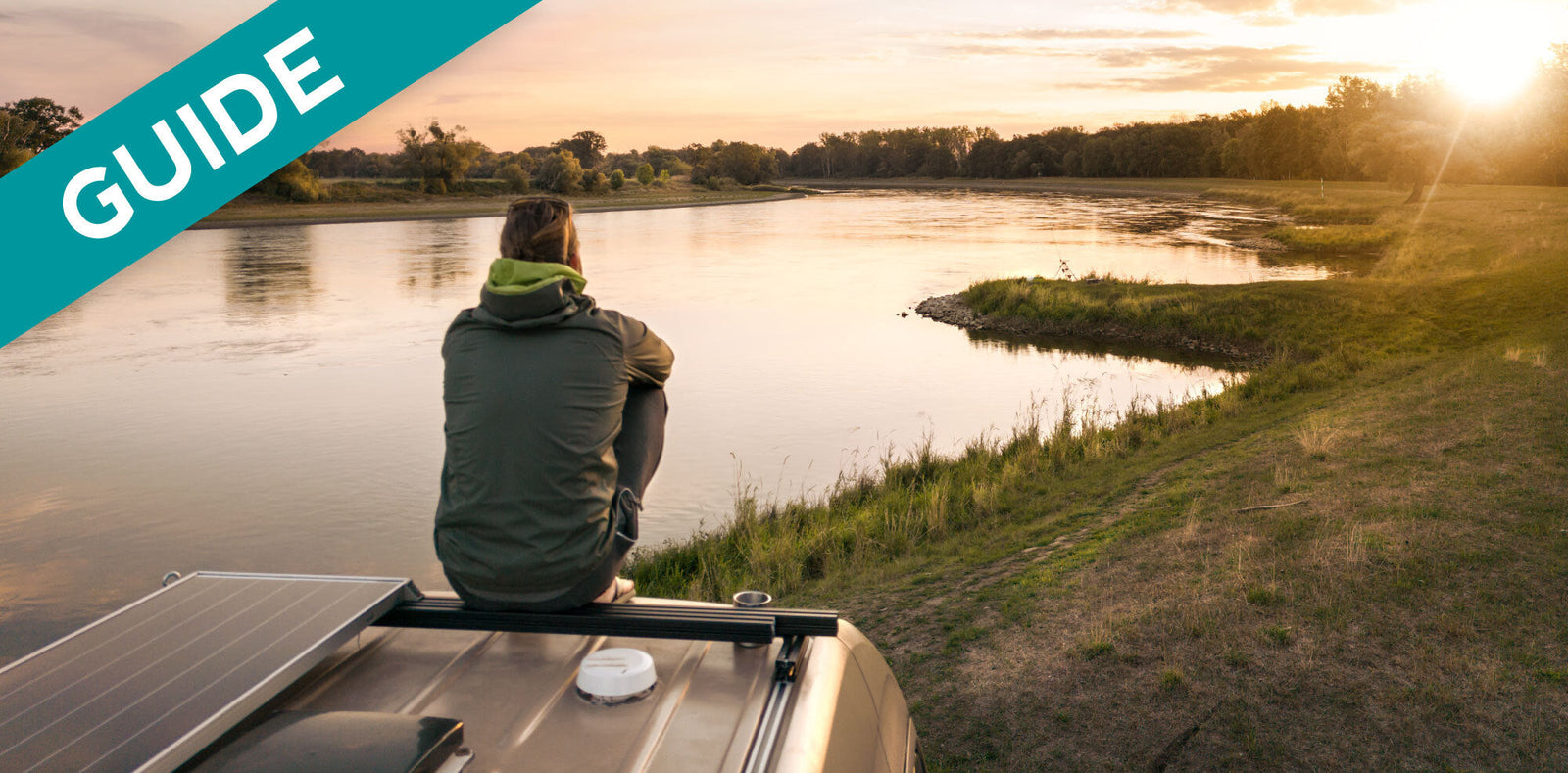 Person sitting on RV rooftop solar panel, looking at river at sunset, a guide.